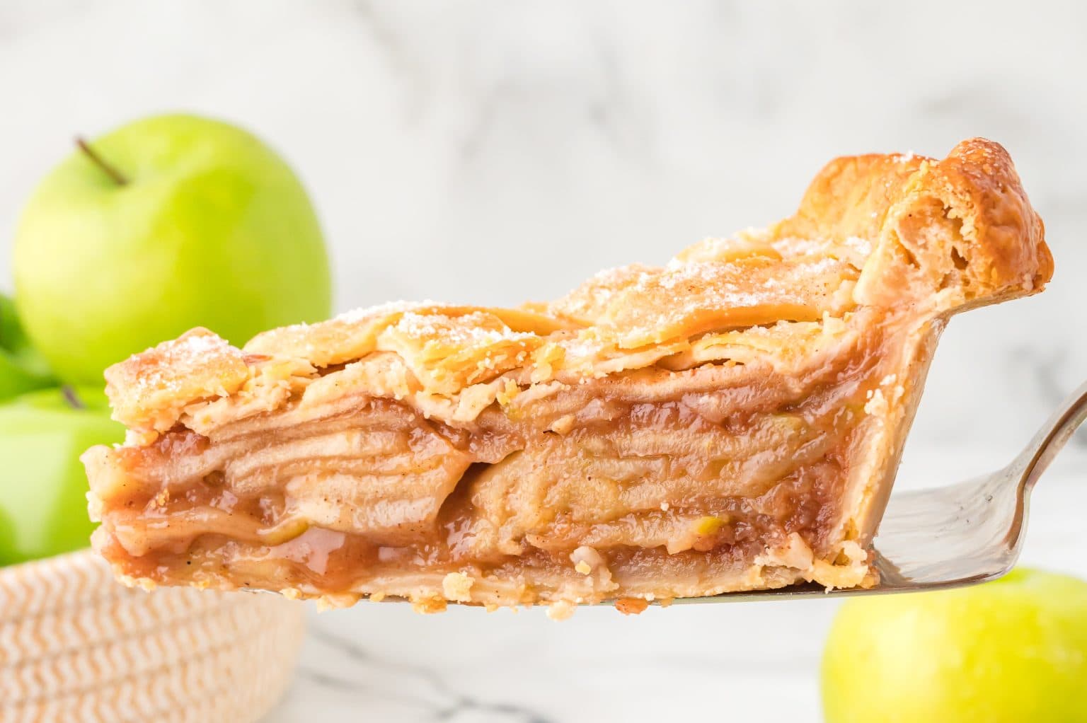 A slice of apple pie, showing layered apples and a golden crust, with fresh green apples in the background.