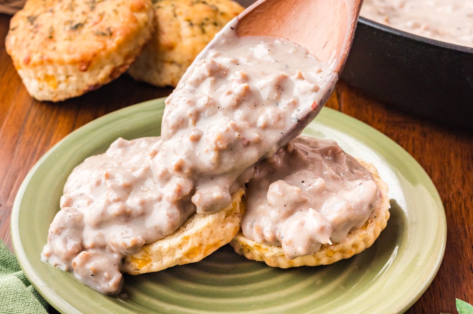 A plate with two freshly baked biscuits is topped with creamy sausage gravy, being spooned over from a wooden spoon. Two additional biscuits are blurred in the background on a wooden table.