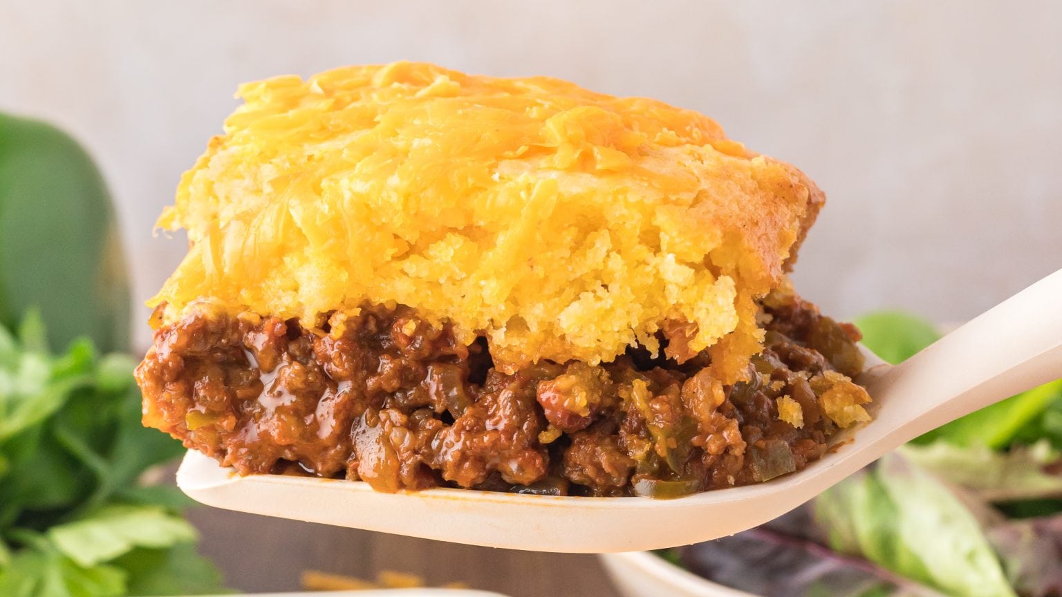 A serving of cornbread casserole topped with melted cheese and a hearty beef filling, held by a wooden spatula against a blurred green backdrop.