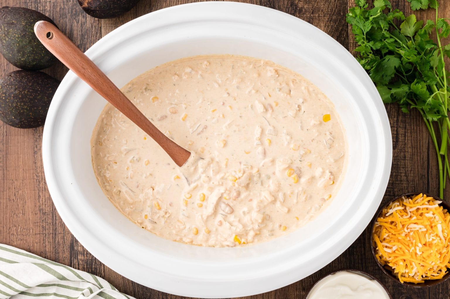 A white crockpot filled with creamy chicken chili, featuring corn and beans, with a wooden spoon inside. Surrounding the crockpot are whole avocados, a bunch of cilantro, a bowl of shredded cheese, and a small bowl of sour cream on a wooden surface.
