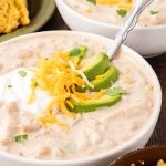 White Chicken Chili in bowls with avocado, cheese and sour cream with a spoon.