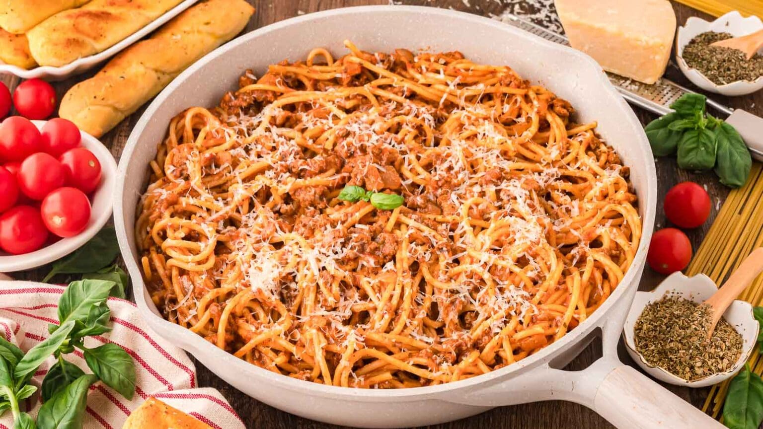 A pan filled with spaghetti bolognese topped with grated cheese and a basil leaf. Surrounding the pan are ingredients like tomatoes, spaghetti, herbs, and a block of cheese on a wooden table. Breadsticks are placed nearby.