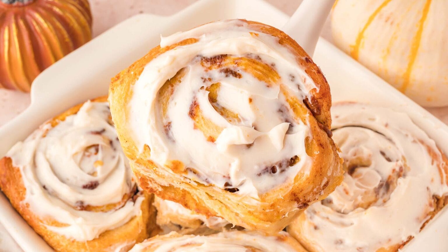 A close-up of a tray of cinnamon rolls topped with creamy icing. One roll is being lifted out, highlighting its spiral shape and glistening glaze. Small pumpkins are seen in the background, suggesting an autumn theme.