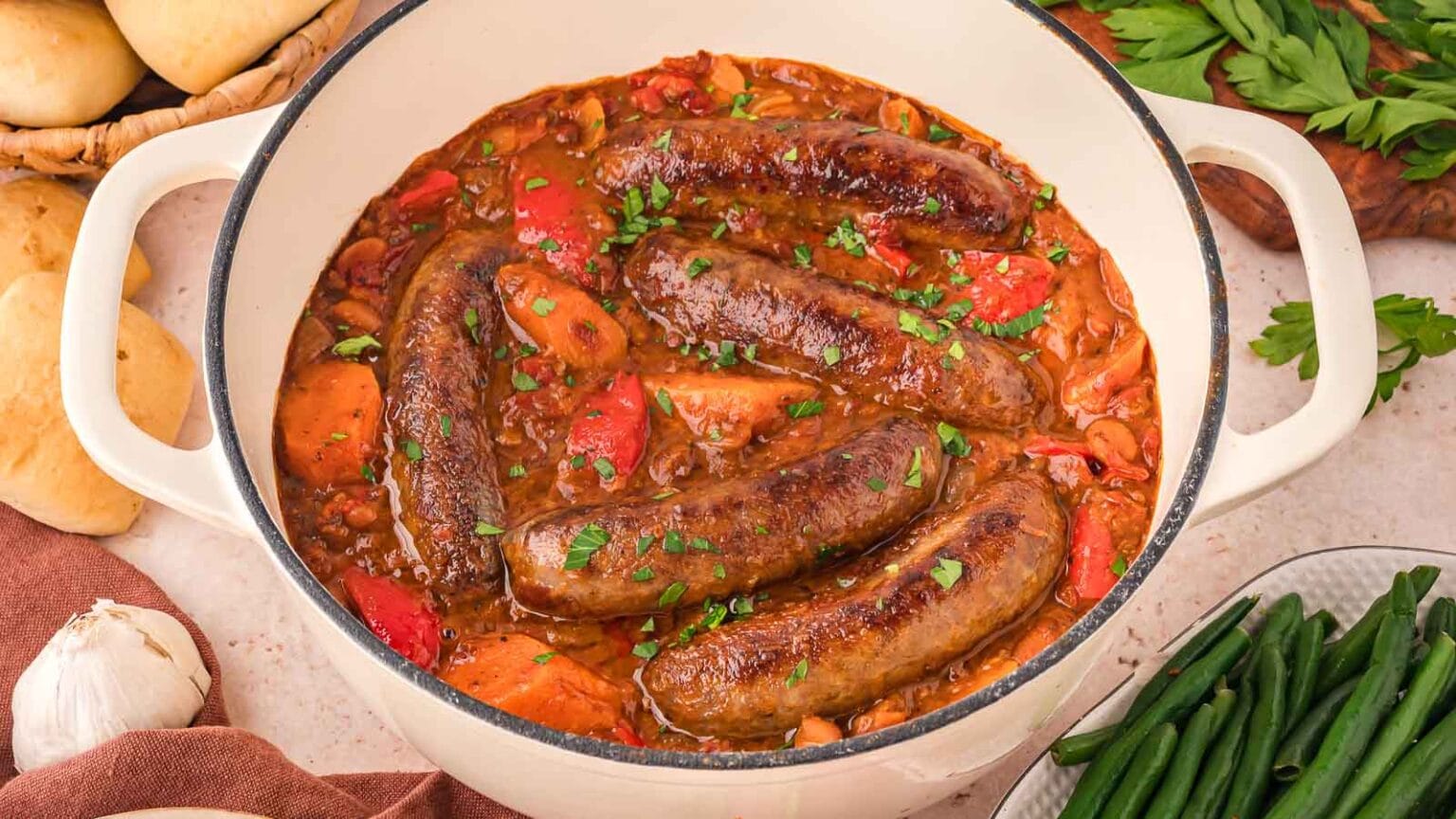 A white pot filled with cooked sausages in a rich tomato stew with carrots and beans, garnished with chopped parsley. Fresh green beans and bread are seen on the side.
