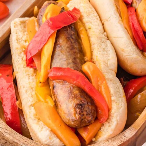 Up close Bratwurst with peppers and onions on hotdog buns on a wooden tray.