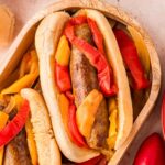 Bratwurst with peppers and onions on hotdog buns on a wooden tray.