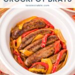 Crockpot brats with peppers and onions in a white slow cooker served with mustard, an easy weeknight dinner by Tessie's Table.