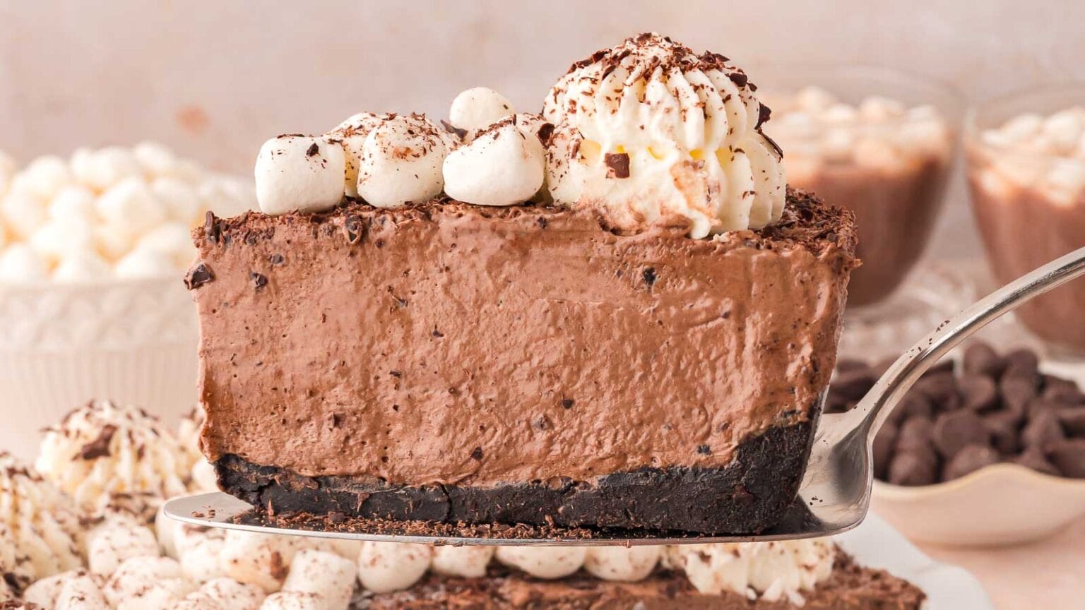 A slice of chocolate mousse cake with an Oreo crust, topped with whipped cream and chocolate shavings, is being lifted from the whole cake. Surrounding the cake are bowls of marshmallows and chocolate chips.