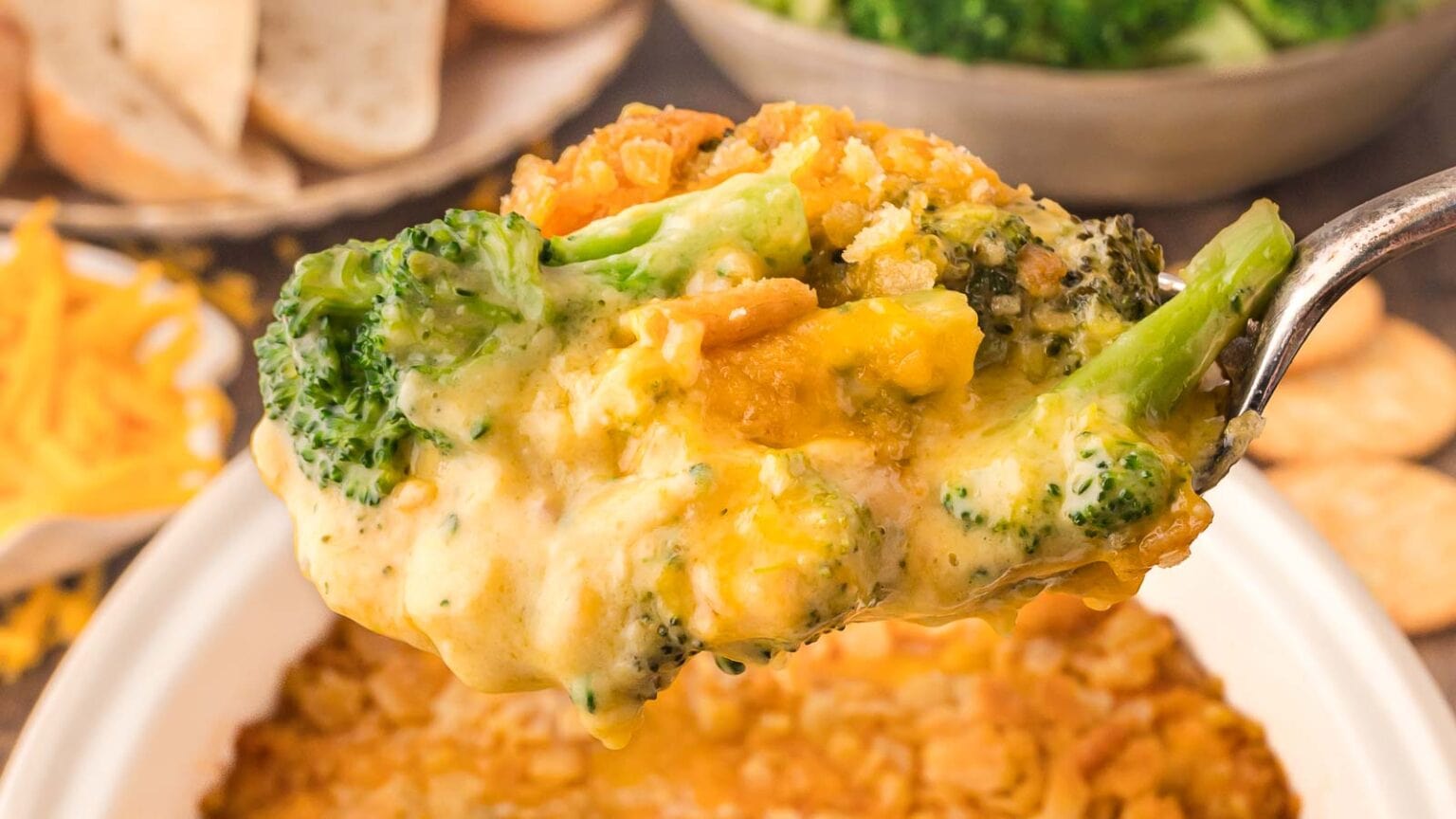 A spoonful of creamy broccoli cheese casserole holds broccoli florets and melted cheese above a baking dish. The background features a bowl of steamed broccoli and slices of bread.