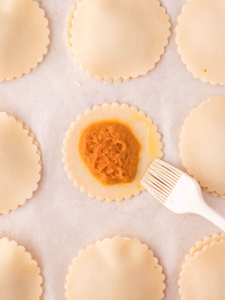 An uncooked hand pie with the filling in the center and an egg wash being brushed around the edges.