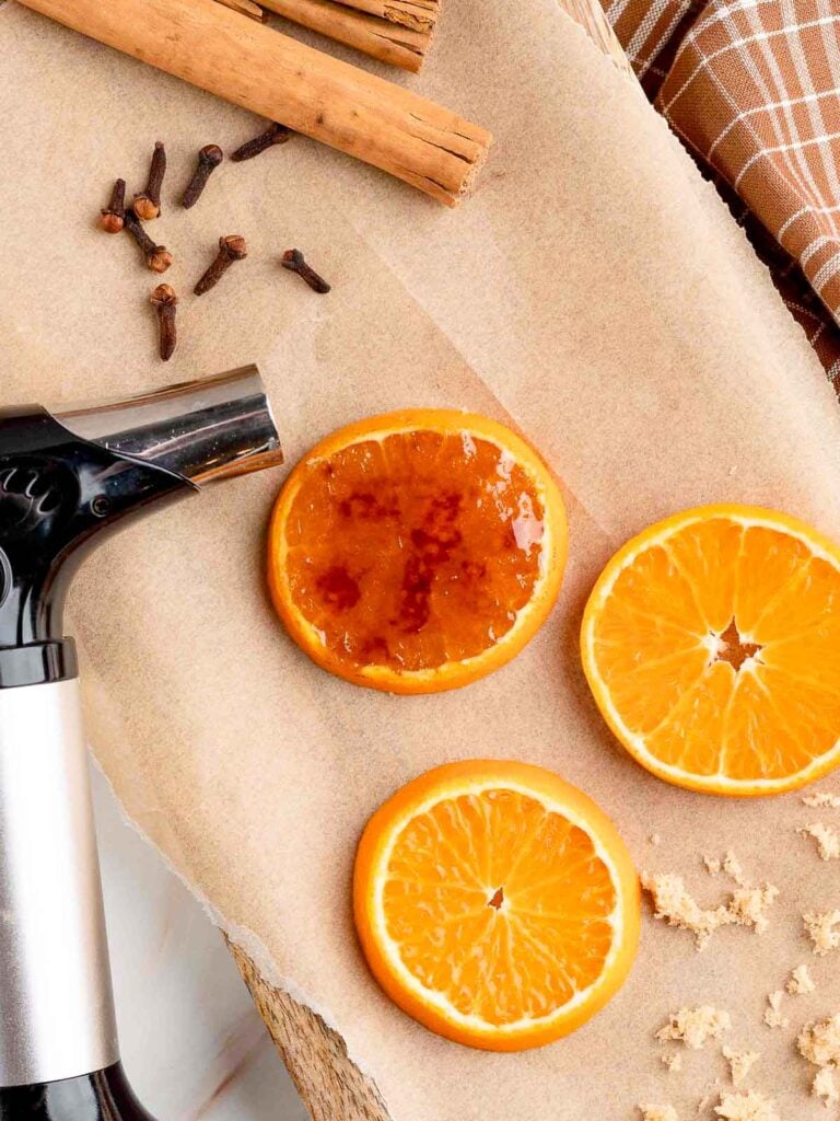 A blowtorch and some sliced of orange on some parchment paper. One slice has been brûléed.