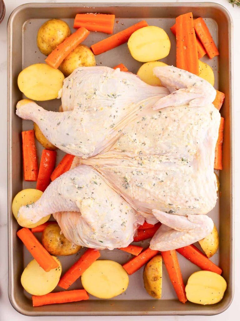 A raw chicken with butter and herbs rubbed into the skin, sitting on top of carrots and potatoes on a baking tray.