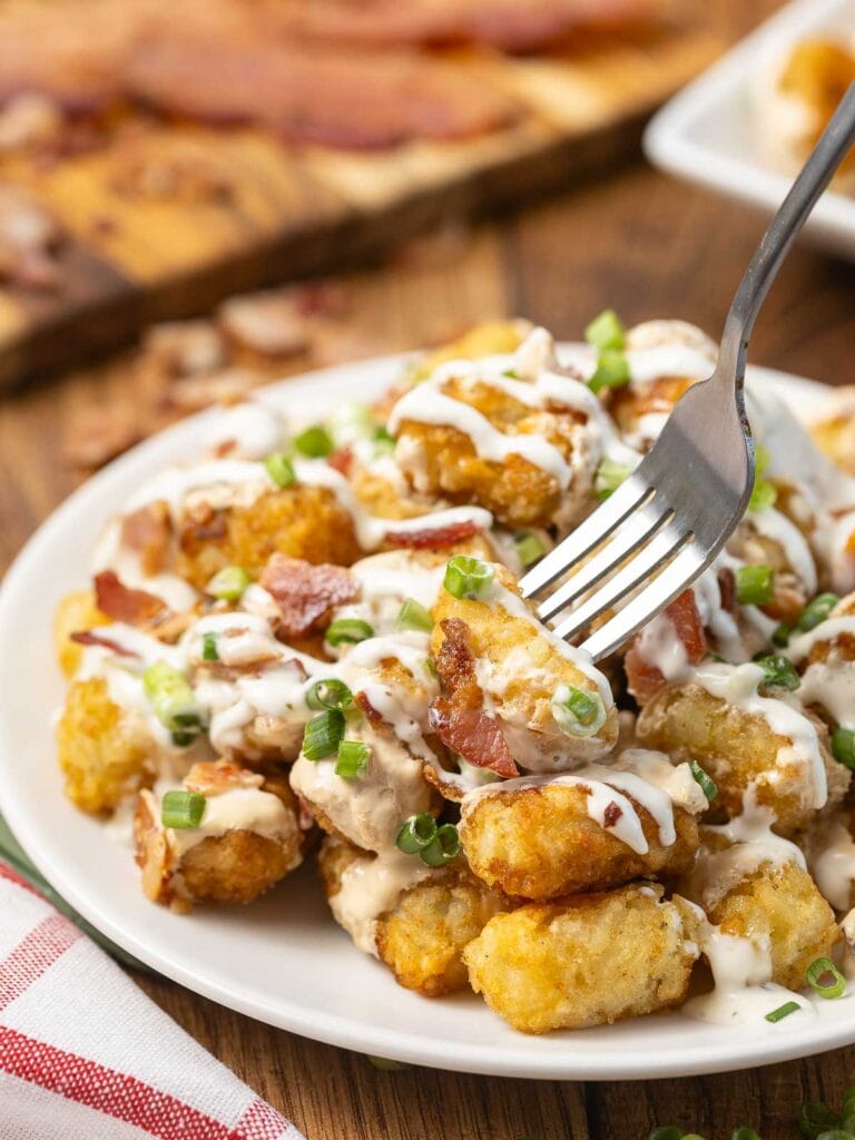 Up close of a fork sticking into a plate of Loaded Tater Tots with queso, bacon and Green onions.