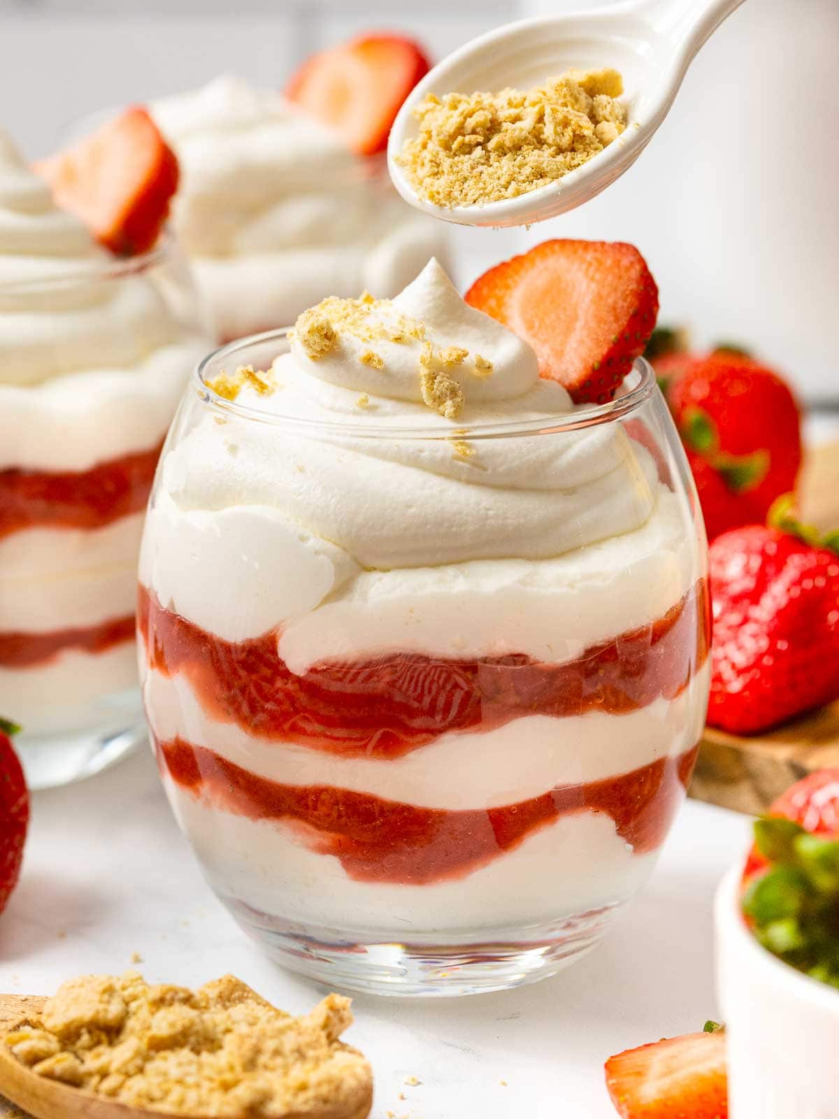 A spoonful of crushed graham crackers being spooned on top of the Strawberries and Cream layers in a glass.