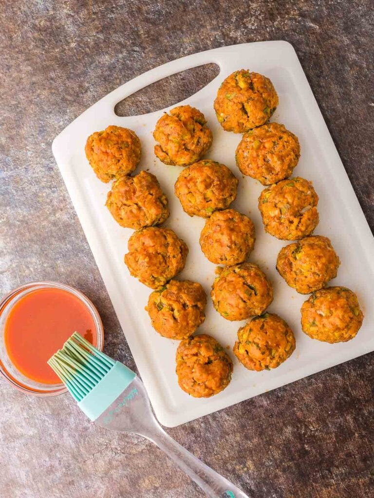 Cooked Buffalo Chicken Meatballs on a white chopping board with some buffalo sauce and a brush beside them.