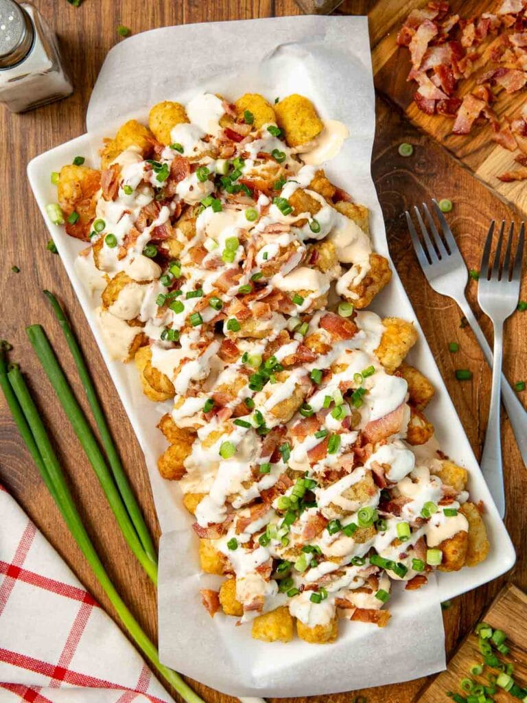 Up close of Loaded Tater Tots with queso, bacon and Green onions on a white serving plate on a wooden table.