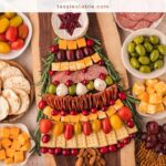 Overhead view of a Christmas tree charcuterie board with a cranberry brie star, rosemary branches and an almond trunk, surrounded by holiday snacks by Tessie's Table.