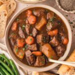 A bowl od show cooker beef stew with fresh bread, mashed potatoes and green beans surrounding it.