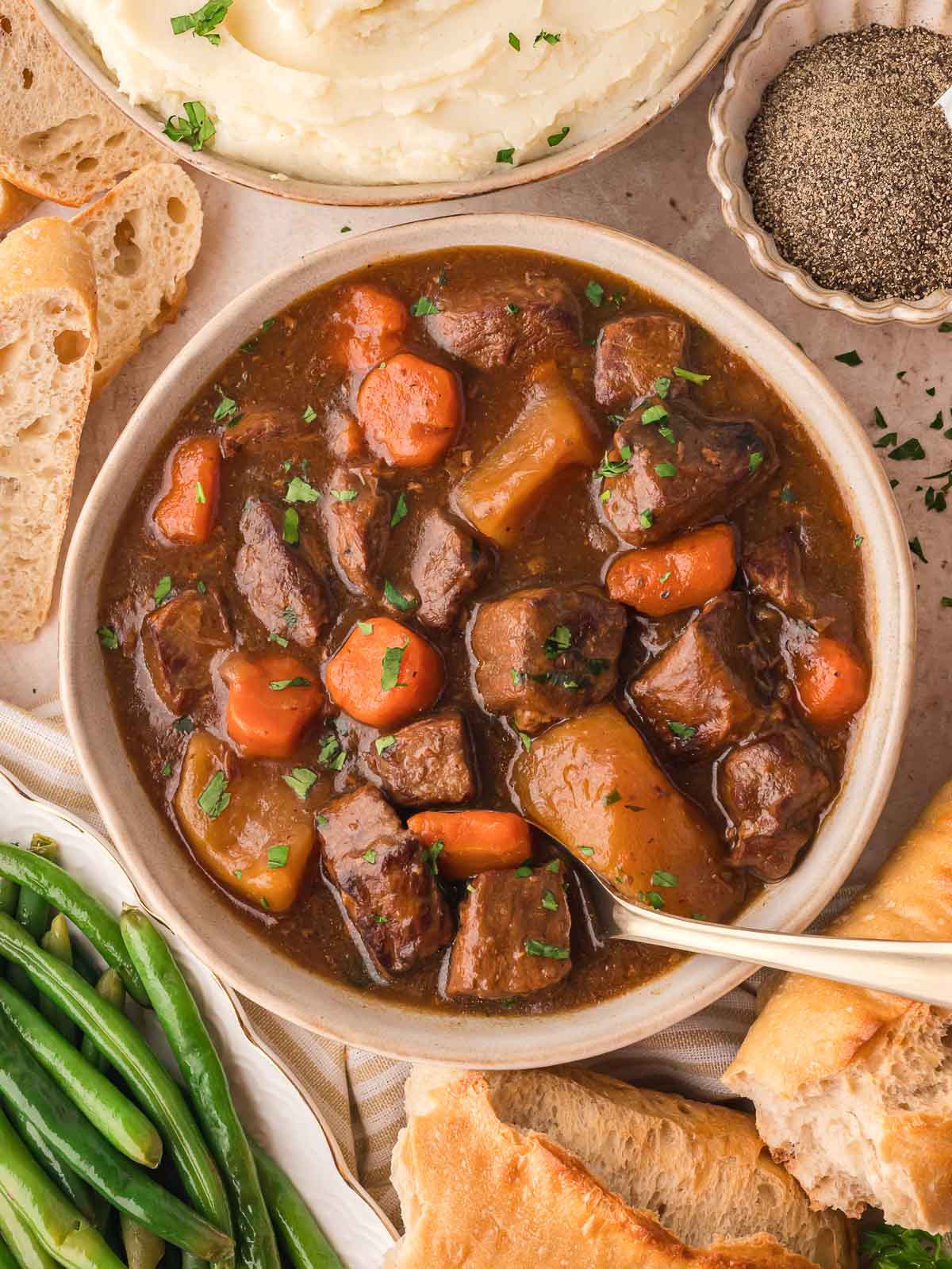 A bowl od show cooker beef stew with fresh bread, mashed potatoes and green beans surrounding it.