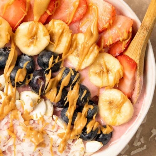 Up close of a smoothie Bowl with blueberries, coconut, banana, Strawberries and peanut butter on top and a wooden spoon.