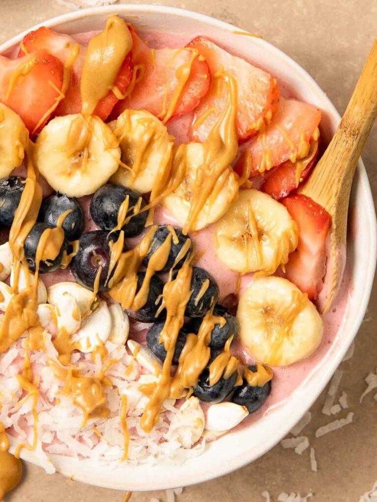 Up close of a smoothie Bowl with blueberries, coconut, banana, Strawberries and peanut butter on top and a wooden spoon.