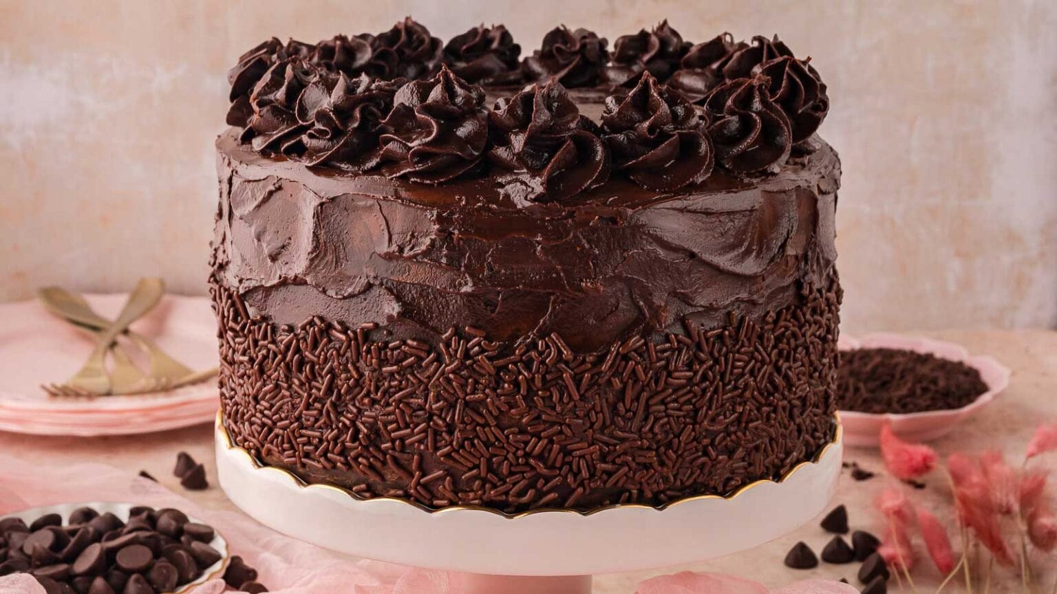 A rich, dark chocolate cake adorned with swirls of frosting and chocolate sprinkles, displayed on a white pedestal against a soft backdrop.