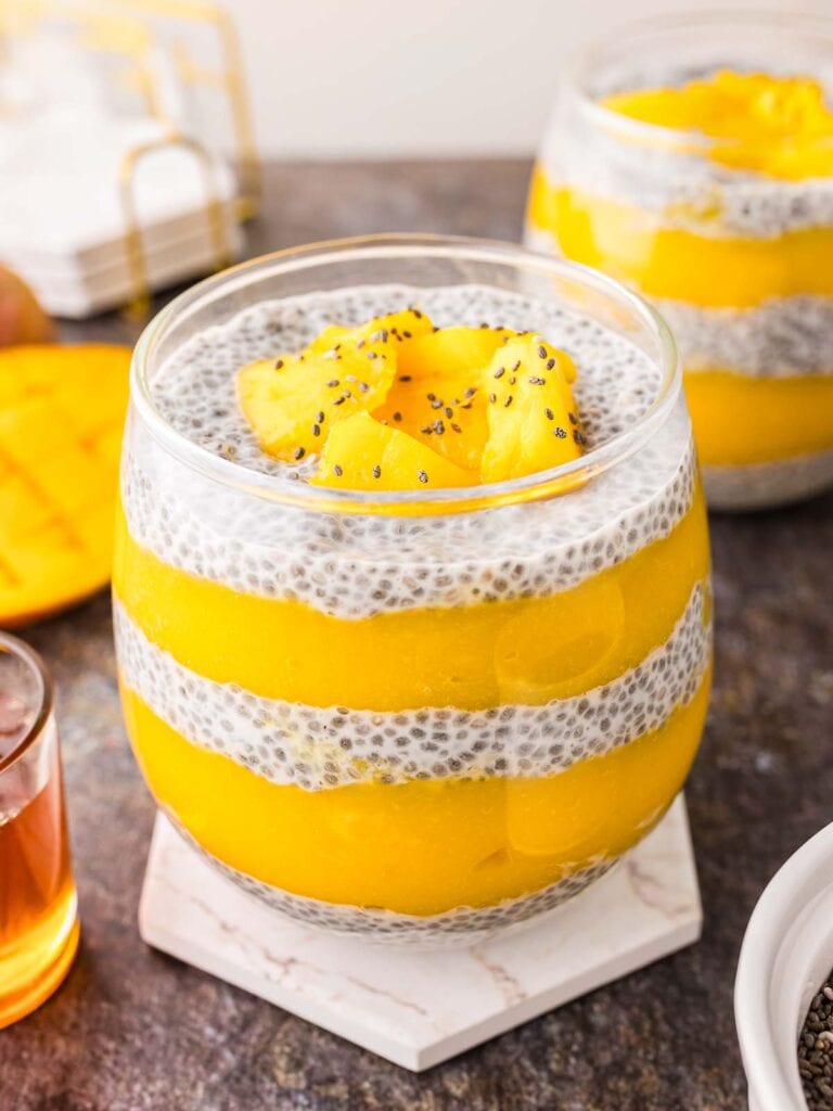 A glass with layers of Mango Chia Pudding sitting on a white coaster.