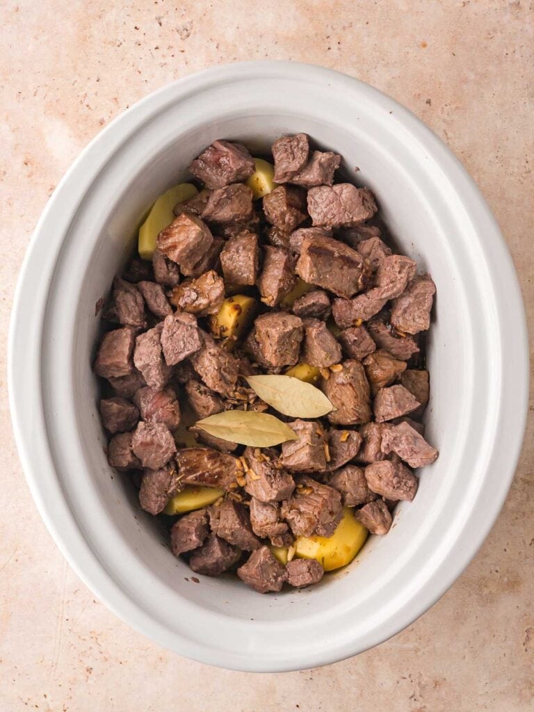 All the ingredients for slow cooker beef stew in a slow cooker with bay leaves on top.