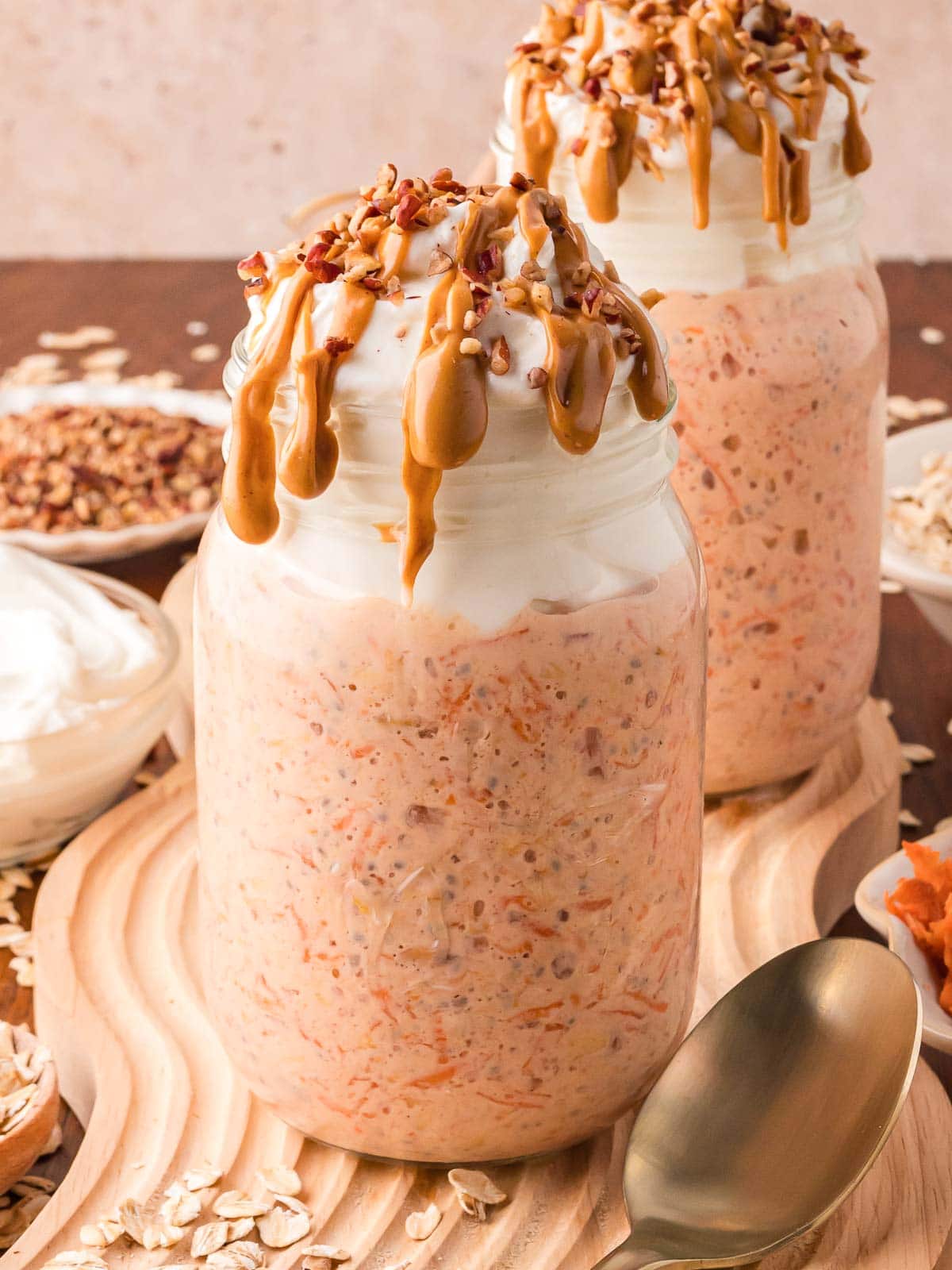 Two mason jars filled with carrot cake overnight oats with greek yogurt on top and peanut butter dripping down the side of the glass.