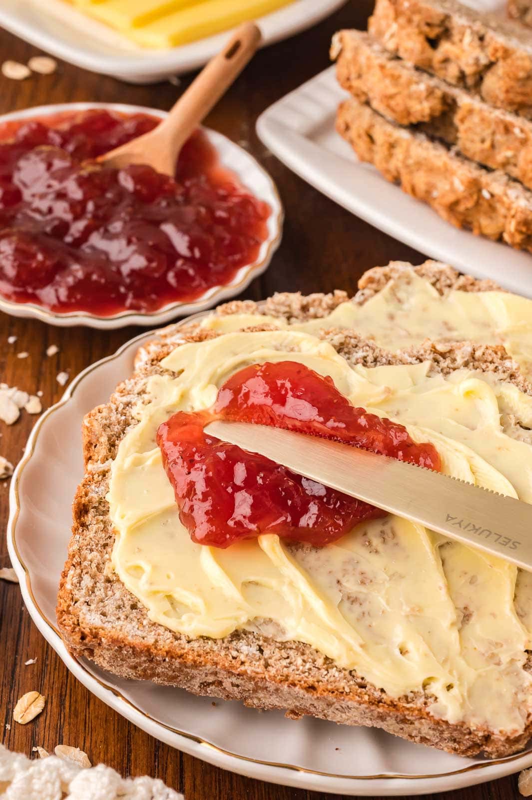 A slice of irish brown bread with butter on it and some jam being spread on top.