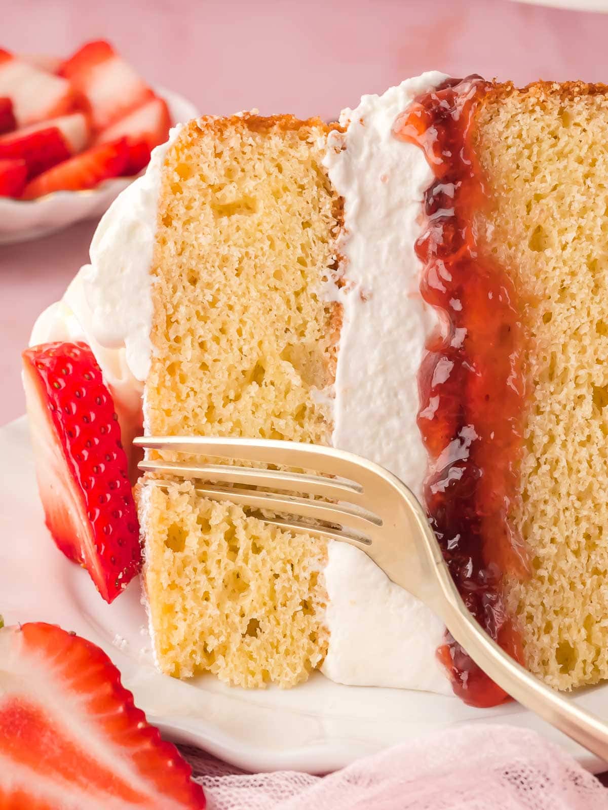 A slice of Victoria Sponge with fresh whipped cream and fresh sliced strawberries and a fork cutting a slice.