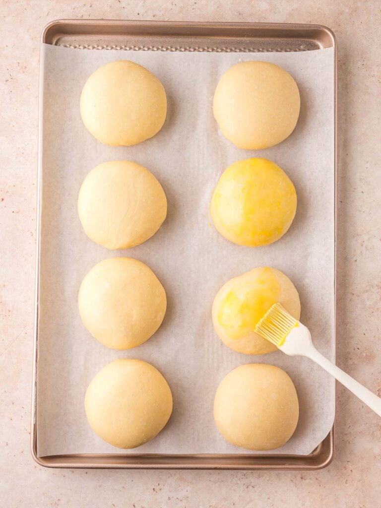 8 Brioche Buns on a baking tray getting egg wash brushed on top.