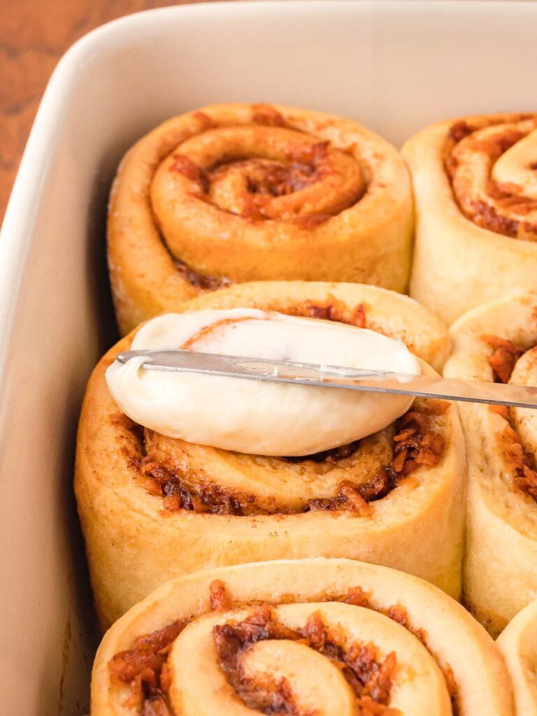 Cream cheese glaze being spread onto carrot cake cinnamon rolls.