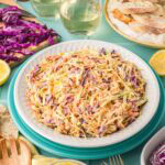 Table setting with a bowl of creamy coleslaw, glasses of white wine, bread, and sliced purple cabbage.