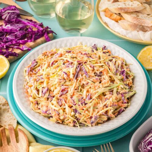 Table setting with a bowl of creamy coleslaw, glasses of white wine, bread, and sliced purple cabbage.