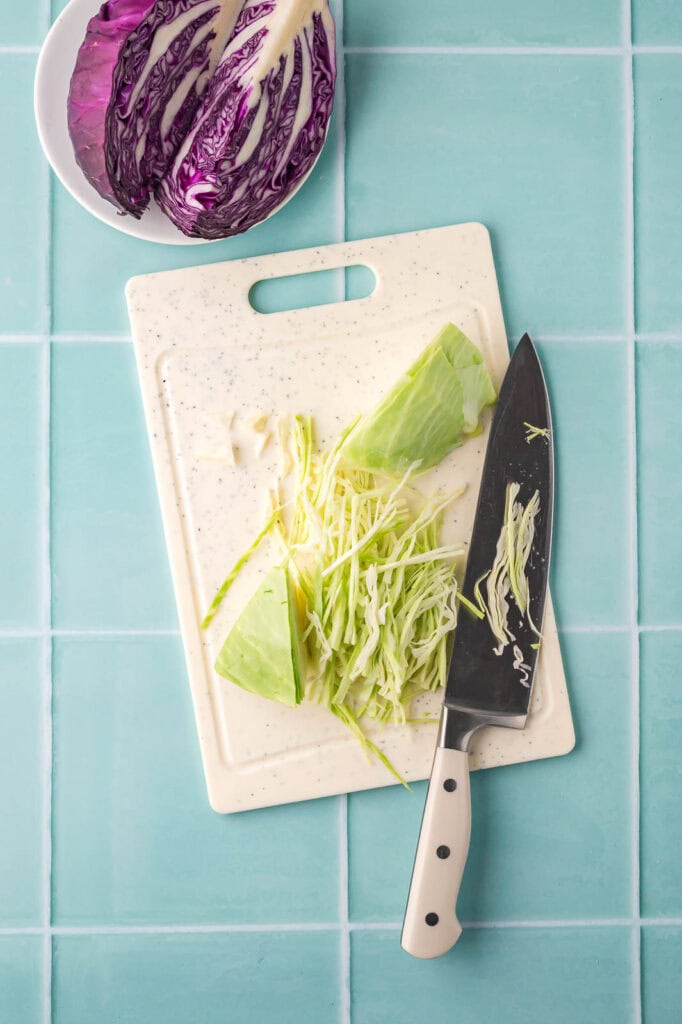 Thinly slicing green cabbage with a knife on a cutting board, purple cabbage shown on the side.