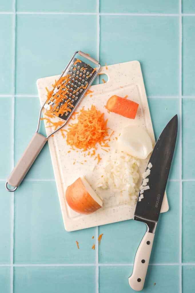 Grating carrots and chopping onion on a cutting board with a knife and box grater.