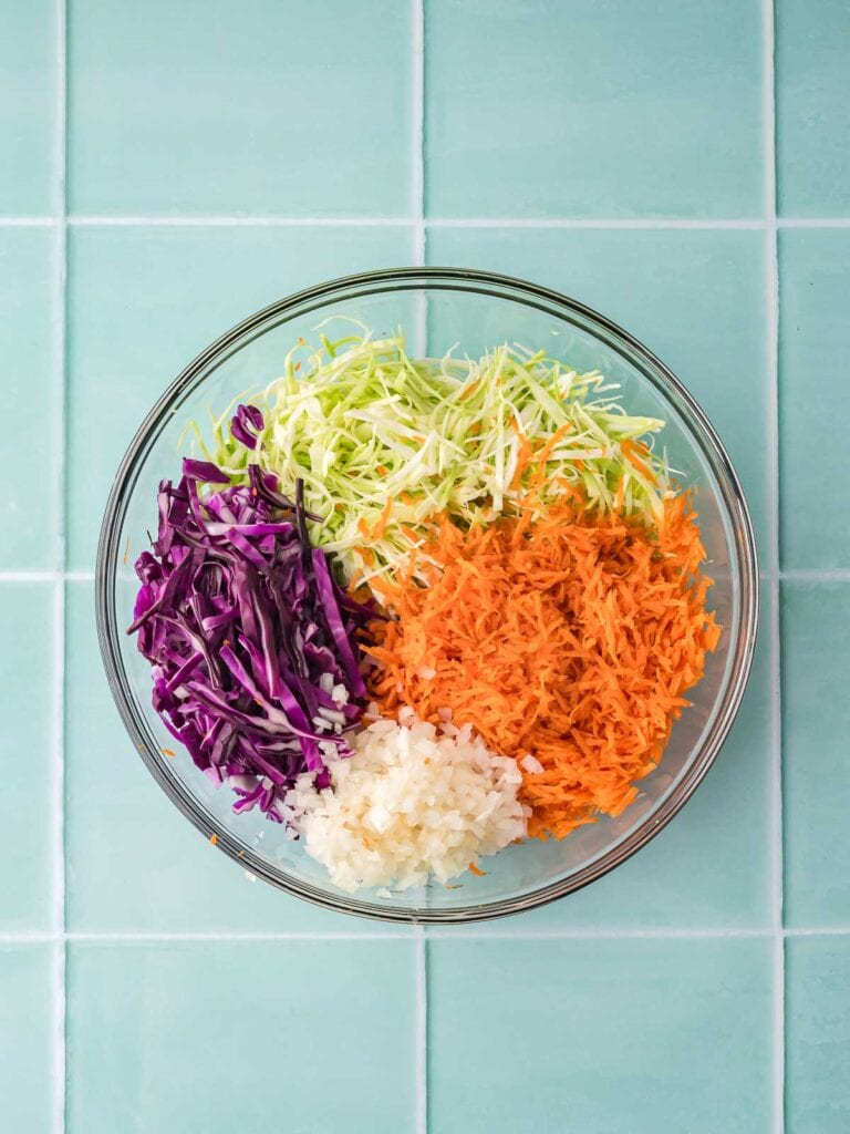 Shredded green cabbage, purple cabbage, carrots, and chopped onion arranged separately in a glass bowl.