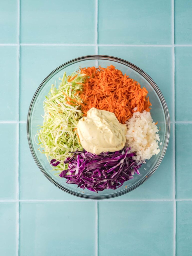 Dressing added on top of shredded cabbage, carrots, and onion in a glass mixing bowl.