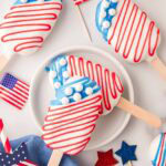 Two red, white, and blue cakesicles stacked on a plate with patriotic props and candy melts in the background