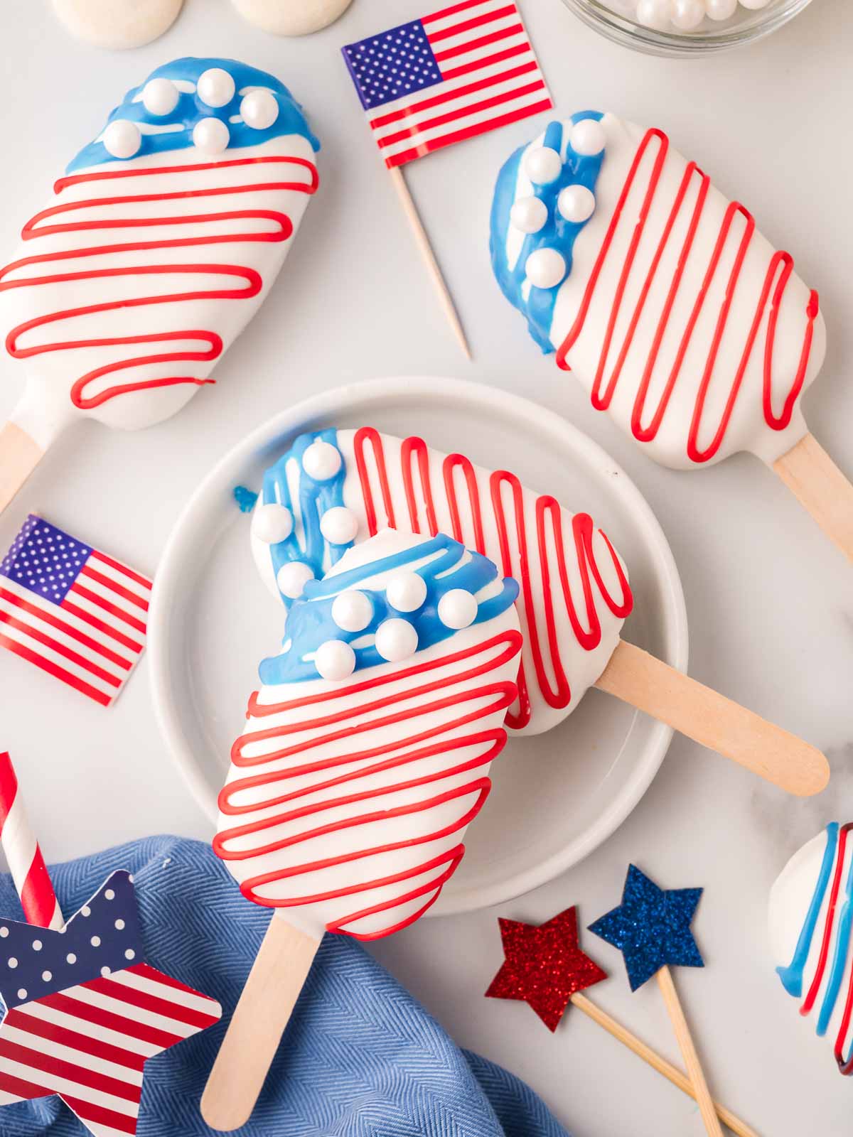 Two red, white, and blue cakesicles stacked on a plate with patriotic props and candy melts in the background