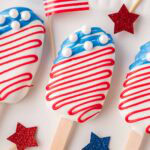 Three American flag cakesicles on a white tray with festive decorations and candy melts nearby