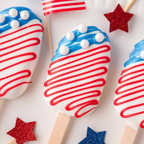 Three American flag cakesicles on a white tray with festive decorations and candy melts nearby