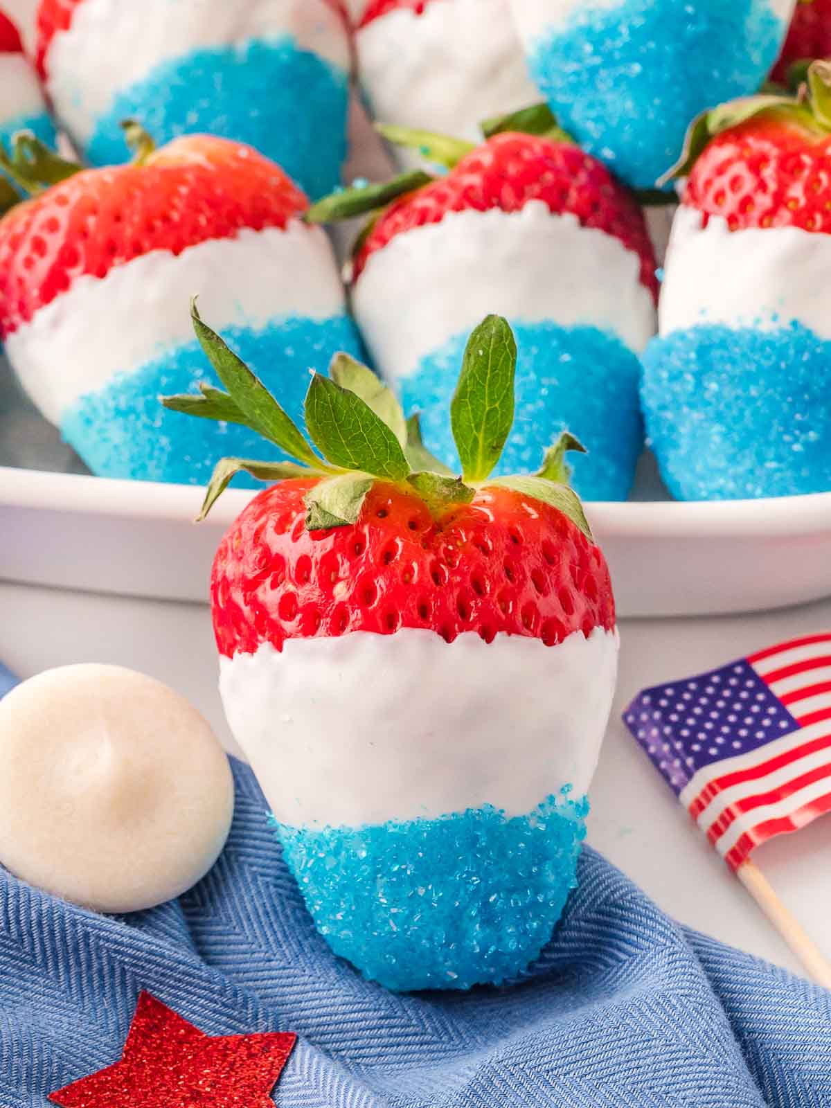 Close-up of a red, white, and blue strawberry dipped in white chocolate and blue sanding sugar with mini American flags in the background.