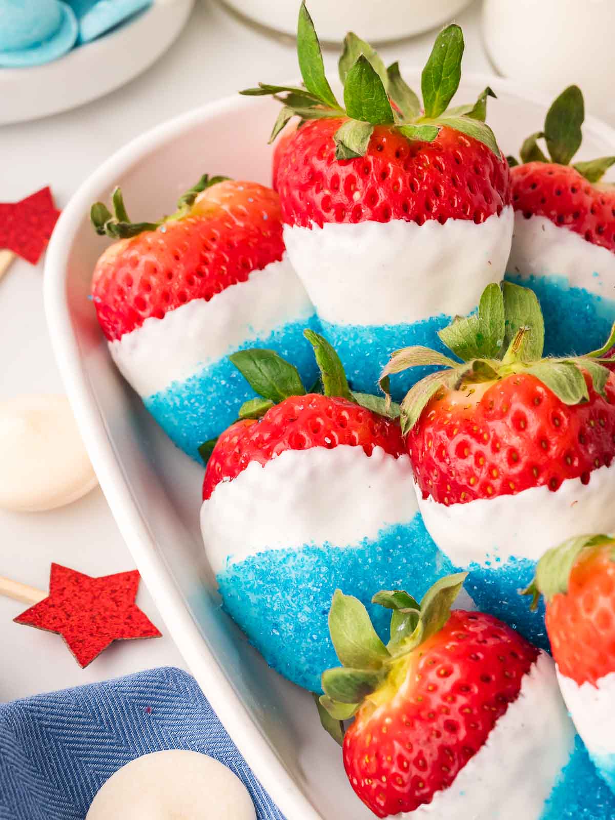 Red, white, and blue strawberries arranged in a white dish, dipped in white candy melts and coated in blue sanding sugar.