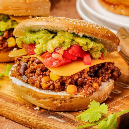 Taco Sloppy Joe on a sesame bun piled high with cheesy taco beef, corn, black beans, tomatoes, and guacamole.