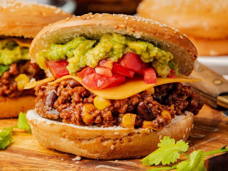 Side angle of a hearty Taco Sloppy Joe filled with ground beef, cheddar, tomatoes, and guac on a wood board.