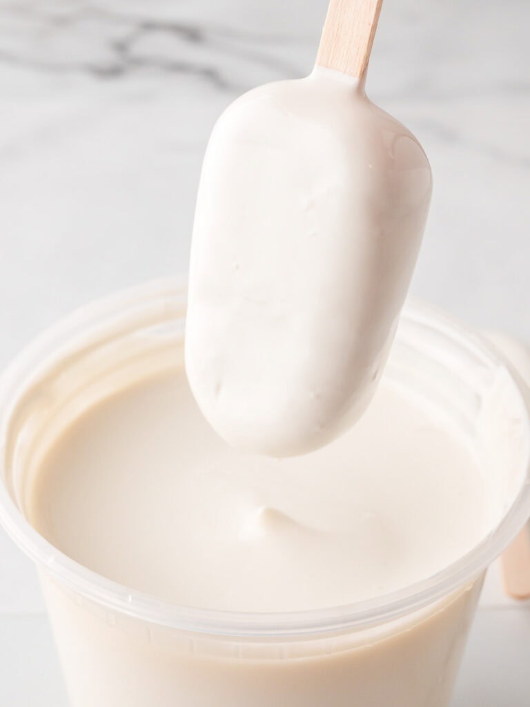 A freshly dipped cakesicle held over a tub of melted white chocolate, showing a smooth even coating.