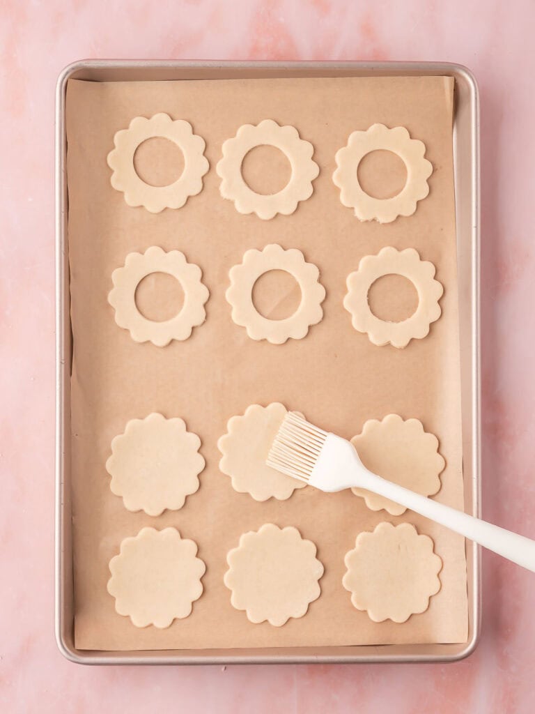 Raw scalloped cookies arranged on a baking tray, with a pastry brush resting on one.