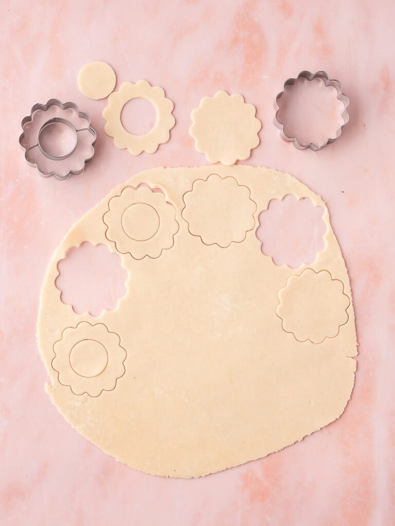 Scalloped cookie dough rounds being cut from pie crust using flower-shaped cookie cutters.